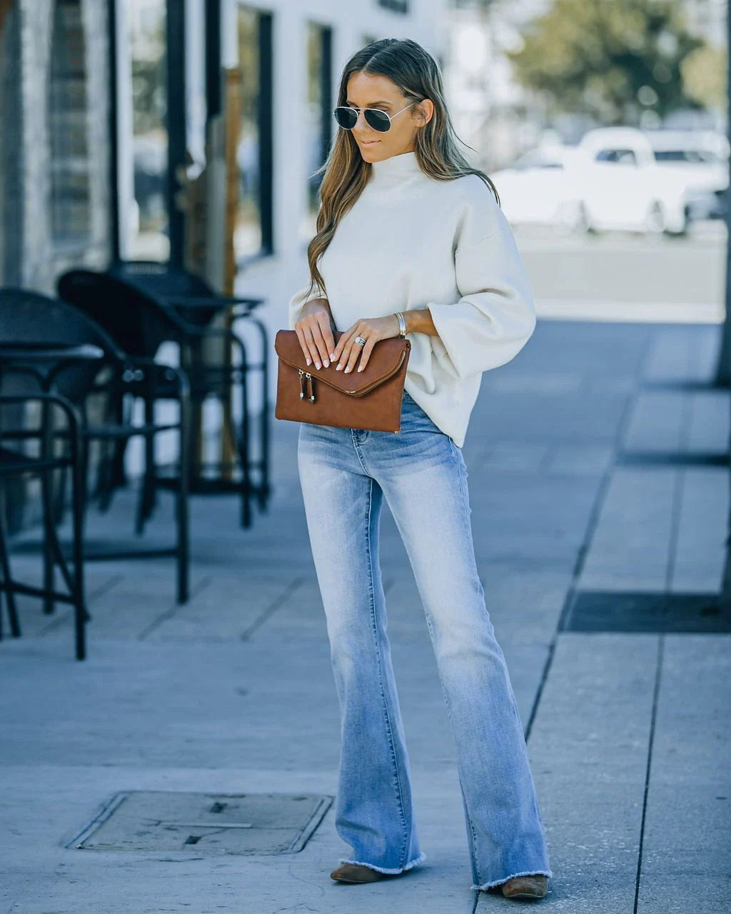 Mujer con pantalón vaquero flare azul claro combinado con un suéter blanco oversize, sosteniendo un clutch de cuero marrón, en una acera urbana