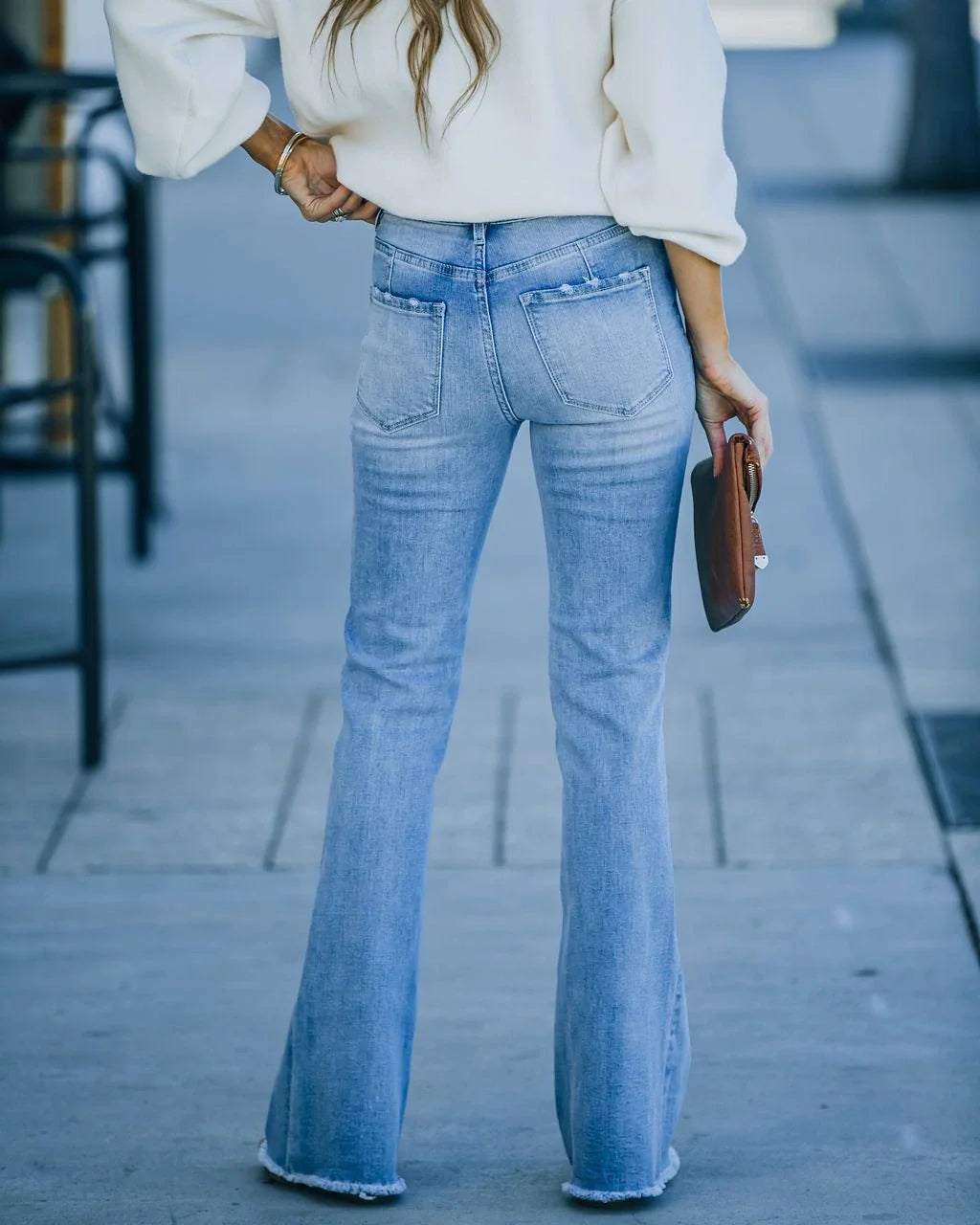 Mujer con pantalón vaquero flare azul claro combinado con un suéter blanco oversize, sosteniendo un clutch de cuero marrón, en una acera urbana